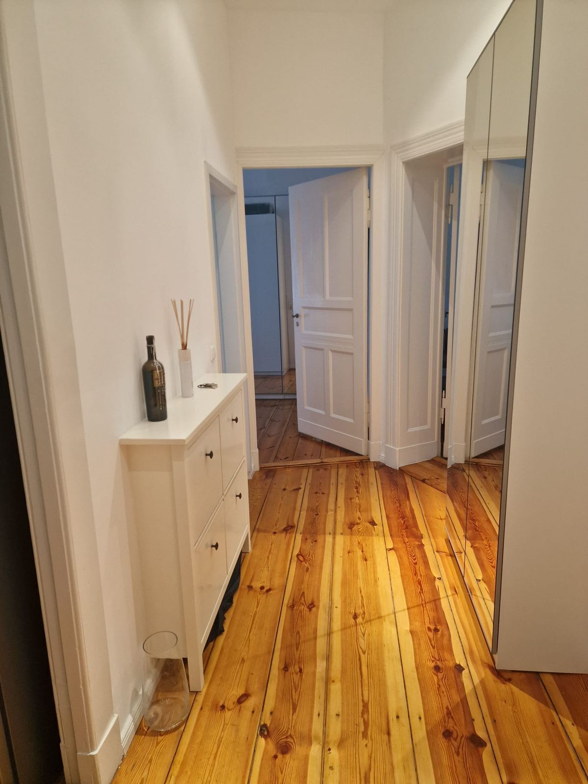 Un pasillo estrecho con suelo de madera, paredes blancas y un espejo a la derecha.