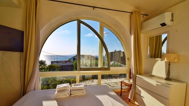 A bedroom with a large arched window, a bed with white linens, a wooden chair, a dresser, and a mirror. - Villa Marghe - Sea View Rooms (Massa Lubrense)