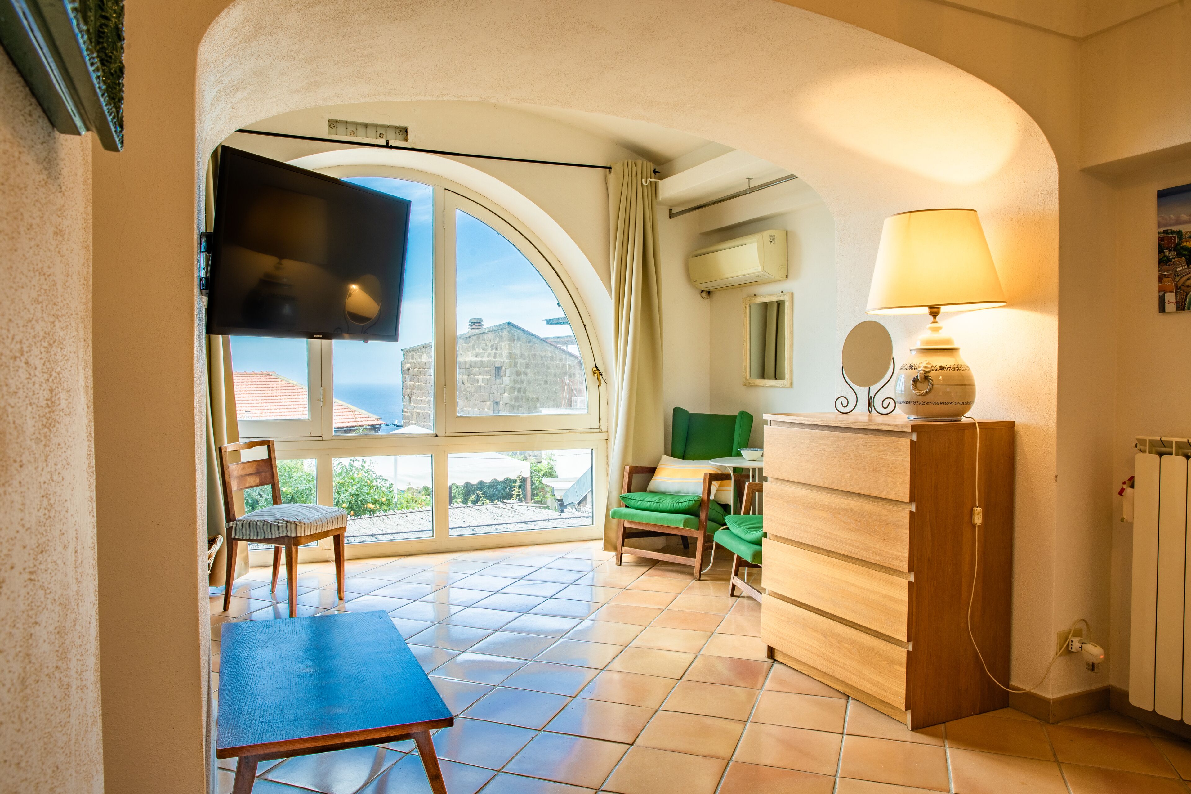 A room with a large arched window, a flat-screen TV, a wooden cabinet, a lamp, and a small table with a chair.
