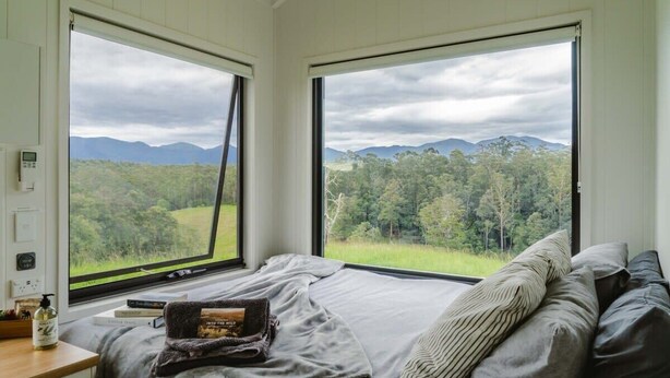 A bedroom with a large bed, two windows with a view of a forest and mountains, a bedside table with a book and a bottle. - Tiny Nancy Summary:<br>H (Bellingen)
