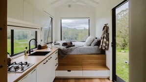 A compact, modern bedroom with a built-in bed, a small kitchenette, and a large window offering a view of the outdoors. - Tiny Nancy Summary:<br>H (Bellingen)