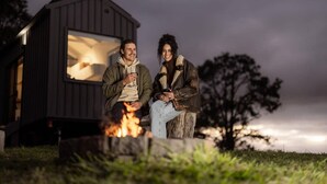 Two people sitting by a campfire in front of a small cabin with a lit interior. - Tiny Nancy Summary:<br>H (Bellingen)