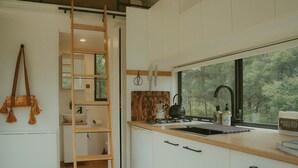 A modern kitchen with a built-in stove, a window with a view of trees, and a wooden ladder. - Tiny Madeline - Laguna (Markwell)