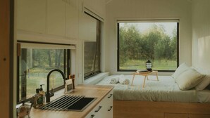 A bedroom with a large window, a bed with pillows, a bedside table with a lantern, and a kitchen area with a sink and coffee machine. - Tiny Madeline - Laguna (Markwell)