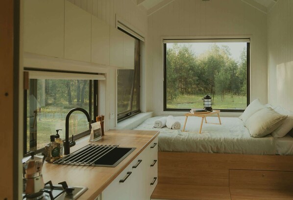 A bedroom with a large window, a bed with pillows, a bedside table with a lantern, and a kitchen area with a sink and coffee machine.