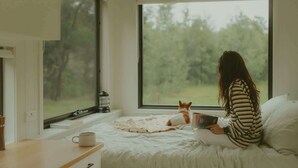 A person sitting on a bed reading a book with a cat beside them, a window with a view of trees, and a bedside table with a cup. - Tiny Madeline - Laguna (Markwell)