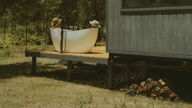 A small wooden cabin with a large window, a dining table with a white bowl and two hats, and a pile of firewood. - Tiny Madeline - Laguna (Markwell)