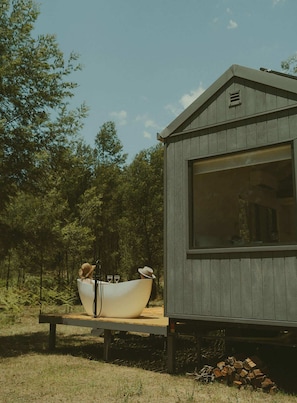 A small wooden cabin with a large window, a dining table with a white bowl and two hats, and a pile of firewood.