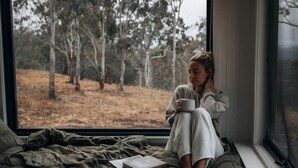 A person sitting on a bed by a window with a book and a cup, overlooking a forested area.