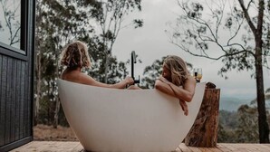 Two people in a large, white bathtub with a view of trees and a wooden structure.