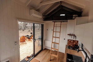 A cabin interior with a wooden deck, a ladder, a small kitchen area, and a sliding glass door leading to the deck.