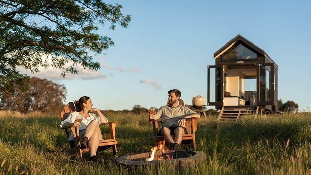 Two people sitting on chairs by a fire pit in a grassy field with a wooden structure in the background. - Tiny Charlotte - Southern Tablelands (Dalton)