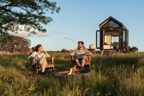 Two people sitting on chairs by a fire pit in a grassy field with a wooden structure in the background. - Tiny Charlotte - Southern Tablelands (Dalton)