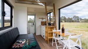 A compact room with a wooden desk, white chairs, a black sofa, and a view of a field through large windows. - Tiny Charlotte - Southern Tablelands (Dalton)