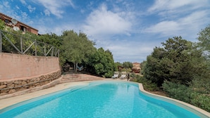 Piscina cristallina con parete in pietra e ringhiera metallica su un lato, circondata da alberi e cielo sereno.