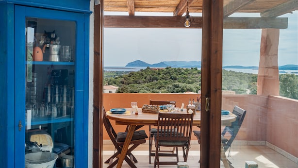 Un balcone con tavolo e sedie in legno, un armadietto blu con vetrate, e vista su un paesaggio montuoso.