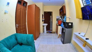 A compact living space with a teal sofa, a kitchen area with a refrigerator, and a TV mounted on the wall. - Lux Suites Ratna Furnished Apartments (Mombasa)