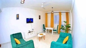 A living room with two green sofas, a small white table, a TV, and a ceiling fan. - Lux Suites Ratna Furnished Apartments (Mombasa)