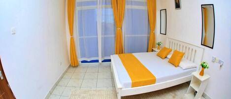 A bedroom with a bed, curtains, a radiator, a small table with a vase, and a mirror on the wall.