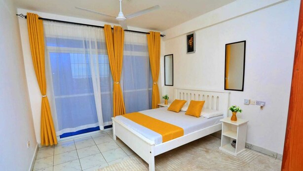 A bedroom with a bed, a nightstand, a ceiling fan, and a window with curtains. - Lux Suites Ratna Furnished Apartments (Mombasa)