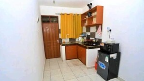 A compact kitchen with a wooden door, a yellow curtain, a black refrigerator, and a tiled floor. - Lux Suites Ratna Furnished Apartments (Mombasa)