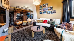 Ein gemütliches Wohnzimmer mit einer Couch, einem Couchtisch und einem Essbereich mit einem Holztisch und Stühlen. An den Wänden befinden sich Bilder, und eine Kristallleuchter ist angebracht.