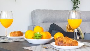 Une table mise pour le petit-déjeuner avec un bol d’oranges et des croissants, deux verres de jus d’orange, et un canapé gris en arrière-plan.
