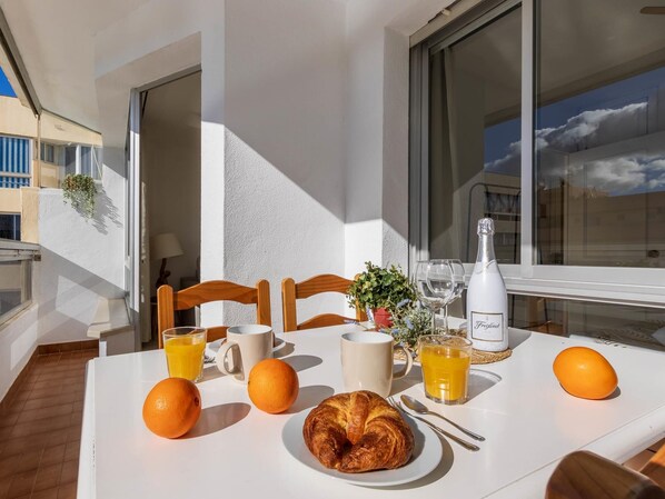 Una tavola per la colazione con succo d'arancia, un croissant e una bottiglia di spumante Freixenet su un balcone con vista sugli edifici.