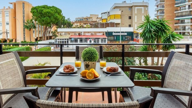 A balcony with a table set for breakfast, including croissants and orange juice, overlooking a cityscape with buildings and a restaurant. - Sol Montemar 23 - Swimming Pool, Parking, 200m from the beach (Torremolinos)
