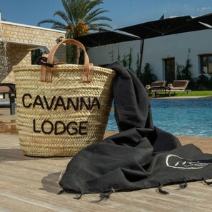 Un cesto intrecciato con la scritta "CAVANNA LODGE", un asciugamano nero e un'area piscina con ombrelloni e palme.