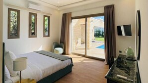 A hotel room with a bed, a desk, a chair, a TV, and a view of a swimming pool. - Cavanna Lodge (Ounagha)