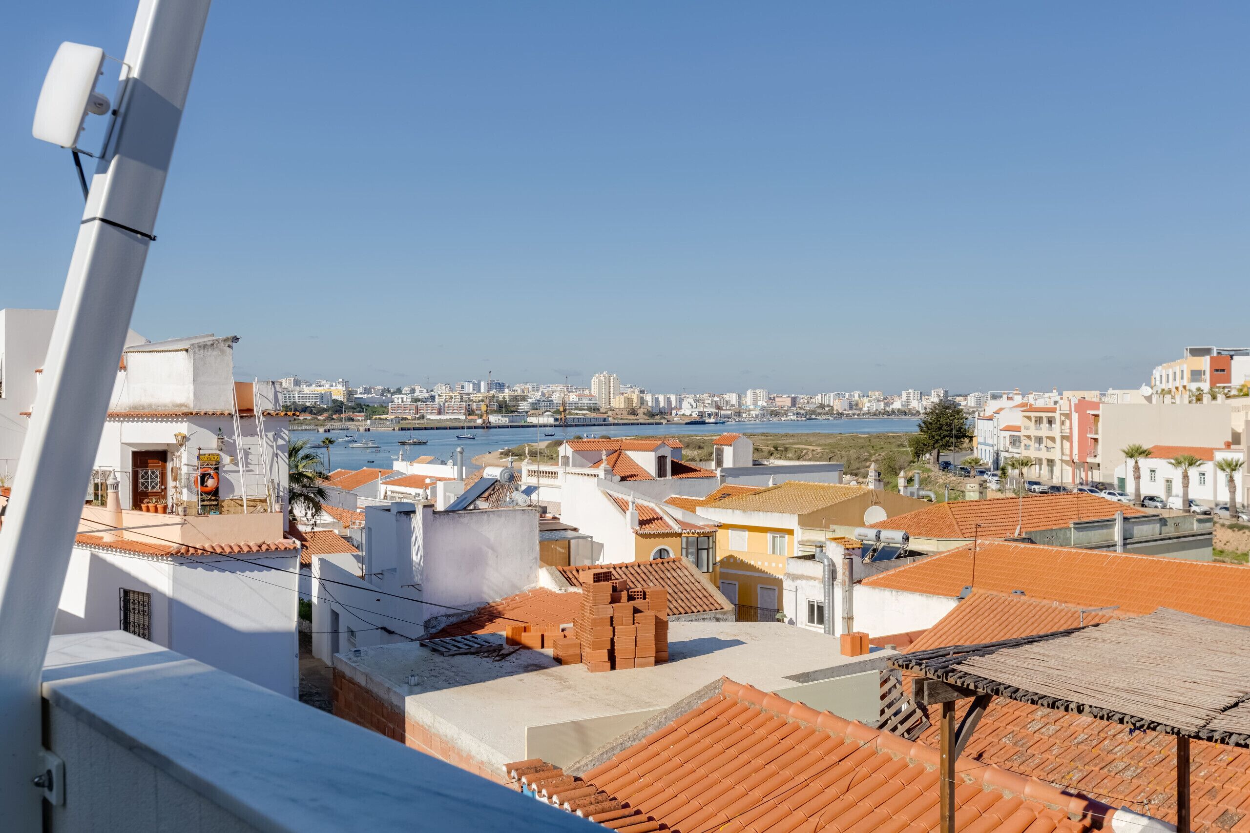 A coastal cityscape with terracotta-roofed buildings, a clear blue sky, and a view of the sea.