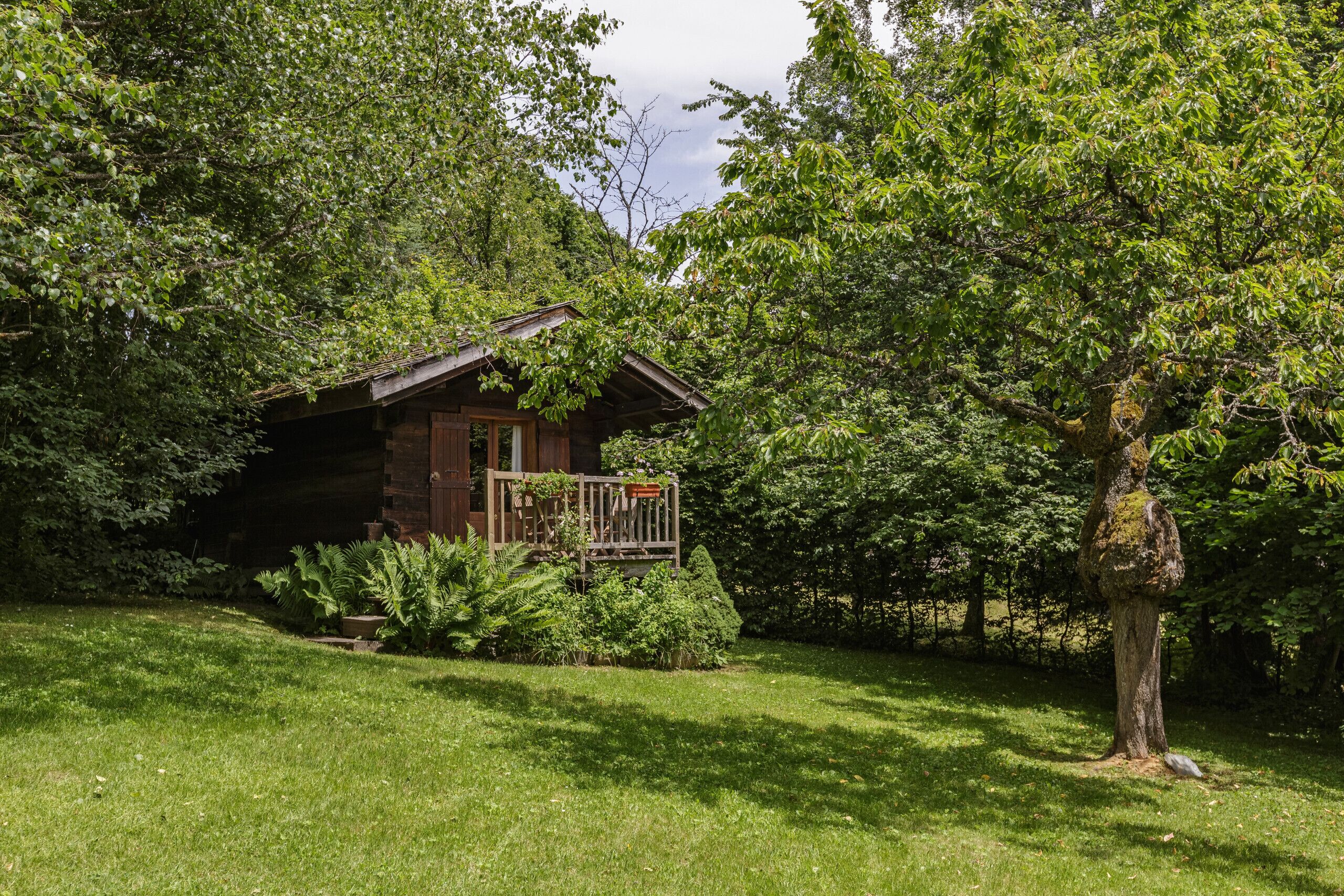 A rustic cabin with a wooden deck, surrounded by lush greenery and trees.