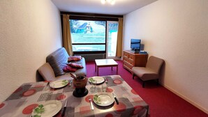 Une chambre meublée avec une table à manger dressée pour deux personnes, un canapé, un fauteuil, un téléviseur et une vue sur une maison par la fenêtre.