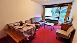 Un salon chaleureux avec une table à manger dressée pour deux personnes, un canapé, une petite table basse et une fenêtre donnant sur l’extérieur.