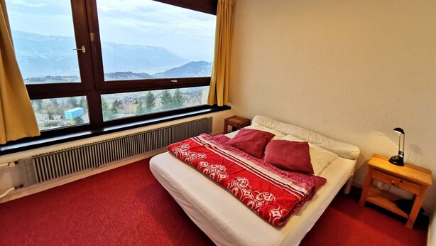 Une chambre avec une grande fenêtre, un lit avec une literie rouge et blanche, une table de chevet avec une lampe et un radiateur.