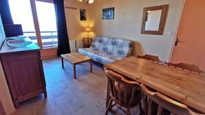 A cozy living room with a wooden dining table, a sofa, and a small coffee table. There is a window with curtains, a lamp, and a ceiling light. - 2-bedroom apt at the foot of the slopes, balcony and elevator (Les Adrets-Prapoutel)