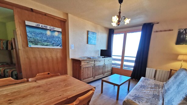 A cozy living room with a wooden dining table, a sofa, and a kitchen area with wooden cabinets and a television. - 2-bedroom apt at the foot of the slopes, balcony and elevator (Les Adrets-Prapoutel)