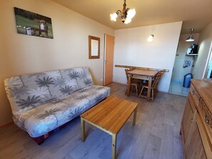 Ein kleiner, gemütlicher Wohnbereich mit einer Couch, einem Holztisch und einem Esstisch mit Stühlen. Eine Deckenleuchte und ein an der Wand befestigtes Regal mit einer Lampe sind vorhanden.