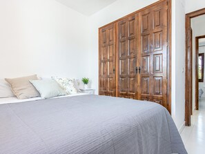 A bedroom with a large bed, a wooden wardrobe, a small bedside table with a plant, and a white wall.