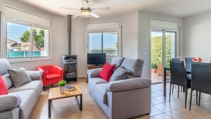 A modern living room with grey sofas, a red chair, a fireplace, a ceiling fan, and a view of the outdoors through large windows. - Beachside Haven with Pool, BBQ, WiFi & A/C – 200m from Sant Pere Pescador Beach (Sant Pere Pescador)