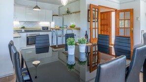 A compact kitchen with white cabinets, a microwave, and a stove. There are two black chairs and a small round table with a bowl. A wooden door with glass panels is visible. - Beachside Haven with Pool, BBQ, WiFi & A/C – 200m from Sant Pere Pescador Beach (Sant Pere Pescador)