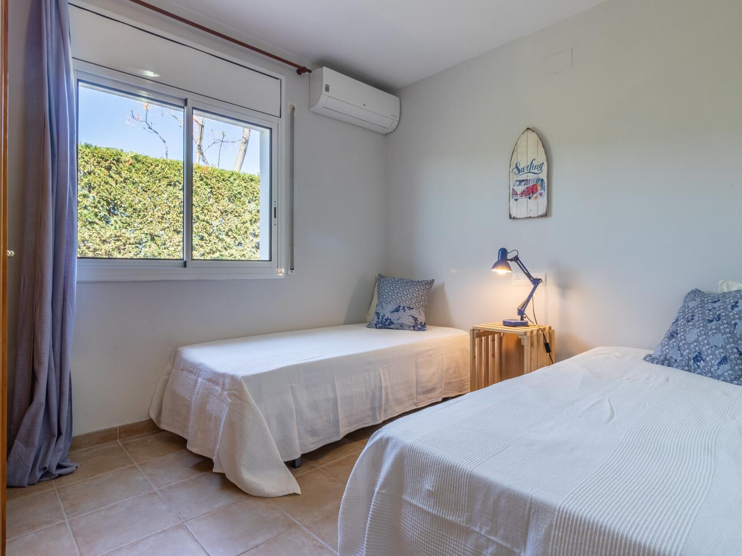 A bedroom with two single beds, a nightstand with a lamp, a surfboard decoration, and a window with curtains.