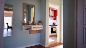 A hallway with a wooden floor, a wall-mounted shelf with decorative items, a mirror, and a door leading to a kitchen with red cabinets. - Apt 6 People, Parking, Air Con, Stunning View (Sant Pere Pescador)
