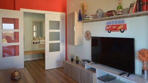 A modern living room with a large flat-screen TV, a wooden entertainment unit, and a bookshelf with books and decorative items. - Apt 6 People, Parking, Air Con, Stunning View (Sant Pere Pescador)