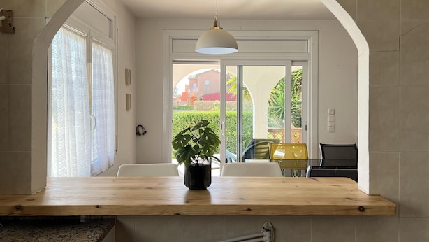 Una cocina con encimera de madera, una planta en maceta, una lámpara colgante y vistas a un espacio exterior con una silla y una mesa a través de una puerta de cristal.
