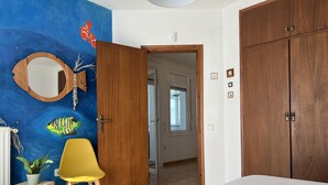 Un dormitorio con una cama, un armario de madera, una silla amarilla, un taburete pequeño, un espejo y una pared decorada con peces pintados.
