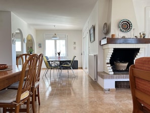 A dining area with a wooden table and chairs, a fireplace with decorative items, and a view of the kitchen through an open door. - Spacious Costa Brava Home: BBQ, WiFi, Parking, Near Beach, Sleeps 7 (Sant Pere Pescador)