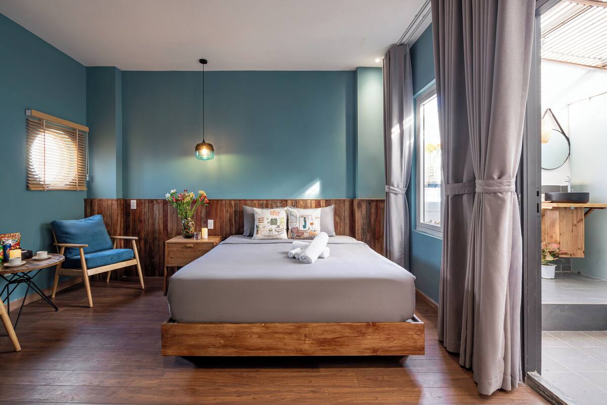 Een moderne slaapkamer met een houten hoofdbord, een groot bed, een blauwe stoel, een kleine tafel en een raam met gordijnen.