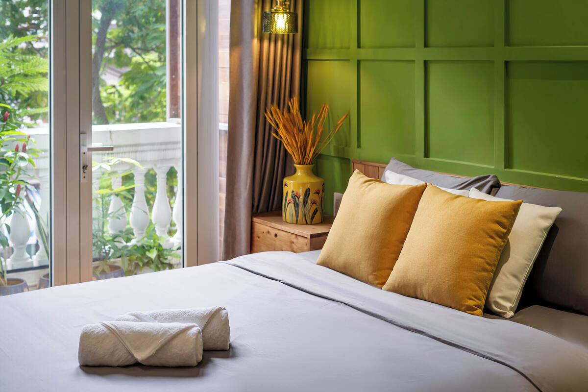 Een opgemaakt bed met twee kussens, een vaas met gedroogde planten, en uitzicht op groen door een raam.